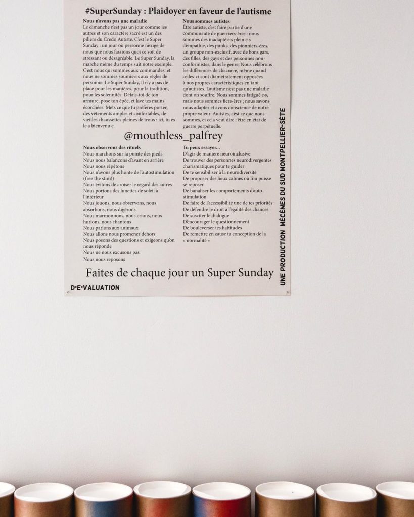 A white wall with a beige poster showing black text written in French. Underneath the poster sits a row of cardboard tubes.