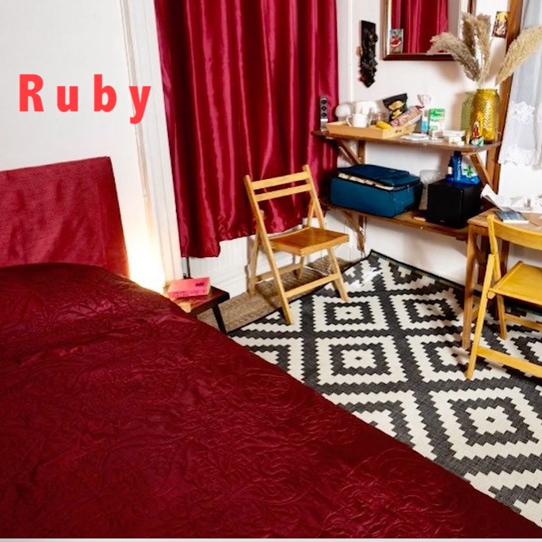 A room with a black and white rug, red curtains and a bed with a red duvet cover. There are wooden chairs and a suitcase in the corner of the room, among other items associated with travelling.