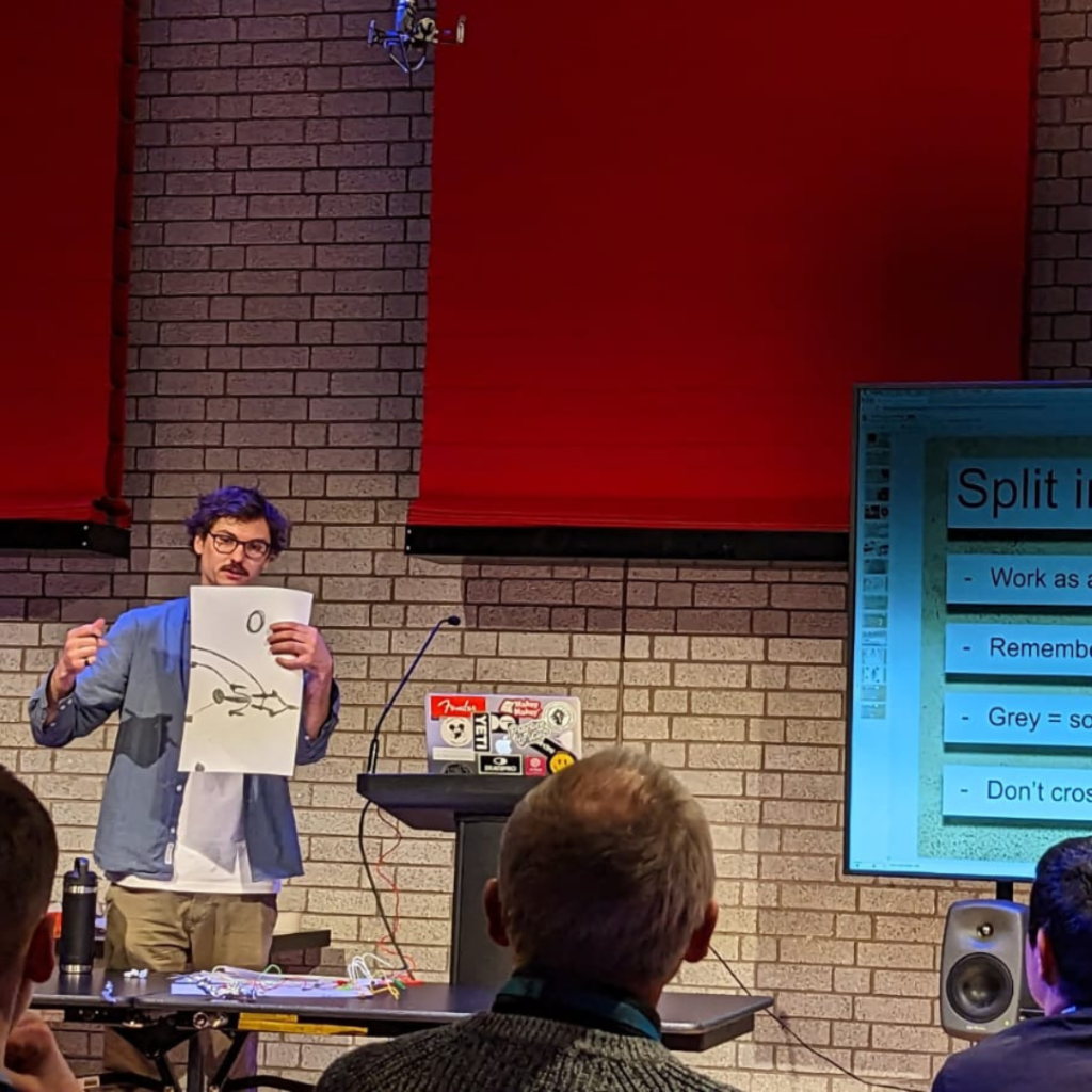 Louis standing in front of a pale brick wall with red fabric hangings. He stands next to a laptop covered in stickers and is holding and pointing towards a piece of paper covered in simple line drawings.