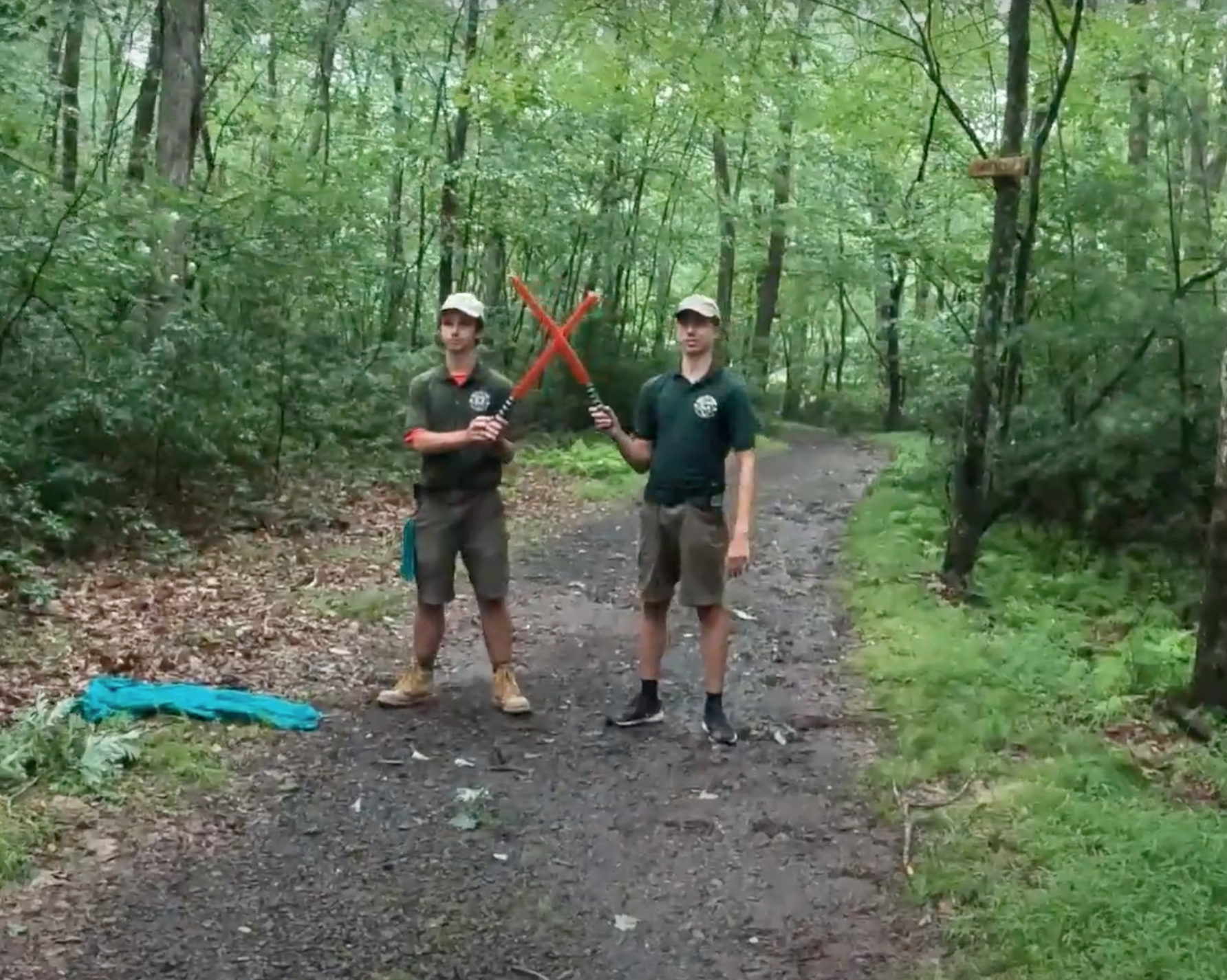 Two Cub Scout leaders standing on a dirt path in a forest. They are holding red toy lightsabers, barring the way forward.