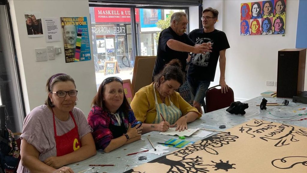 A group of people sit around a table covered in wires and a piece of cardboard. The cardboard has several designs painted in black. Some people are standing to the side of the room.