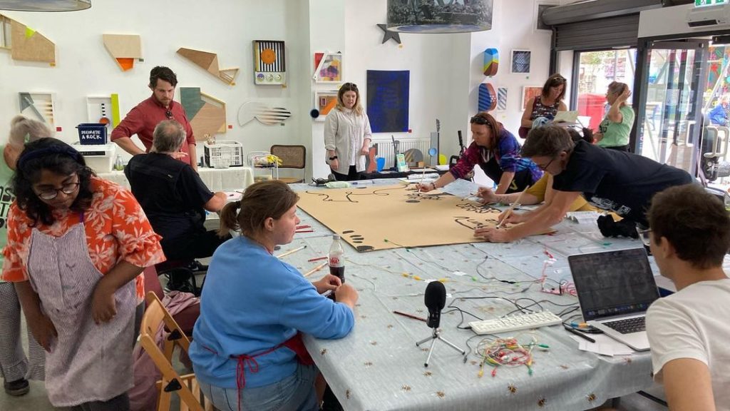 A group of people sit around a table covered in wires and a piece of cardboard. The cardboard has several designs painted in black. Some people are standing.