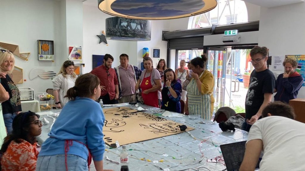 A group of people stand around a table covered in wires and a piece of cardboard. The cardboard has several designs painted in black.