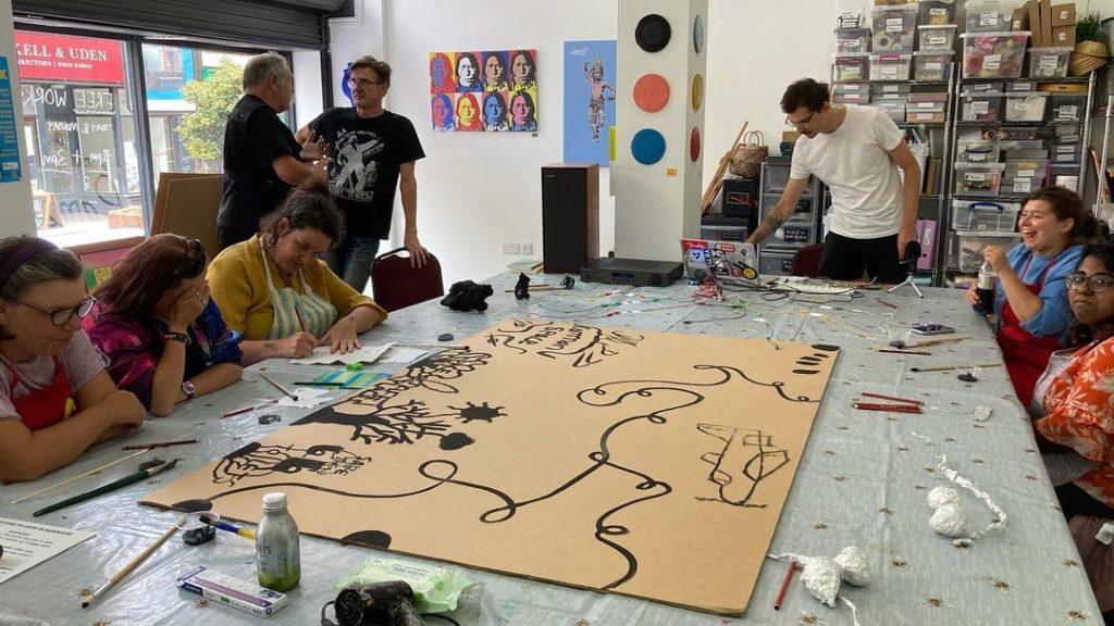 A group of people sit around a table covered in wires and a piece of cardboard. The cardboard has several designs painted in black. Some people are standing to the side of the room.