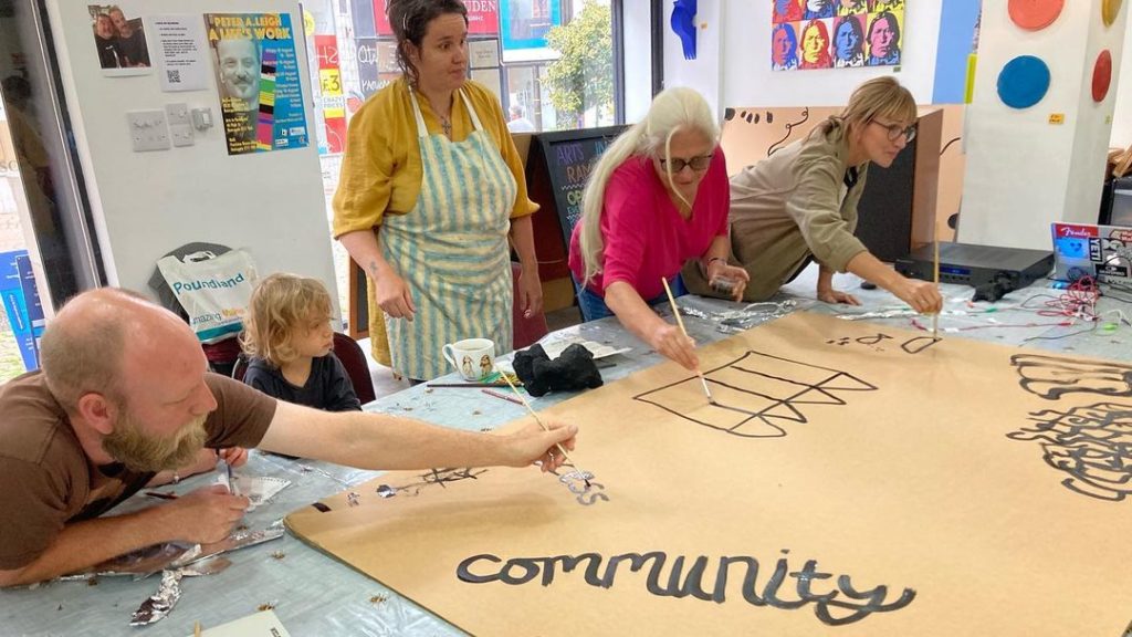 A group of people standing around a table. They are leaning over, painting with black paint onto a piece of cardboard. The word 'community' has been painted by someone at the bottom of the cardboard.