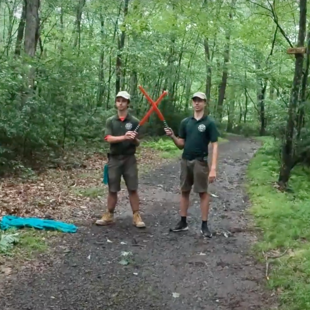 Two Cub Scout leaders standing on a dirt path in a forest. They are holding red toy lightsabers, barring the way forward.