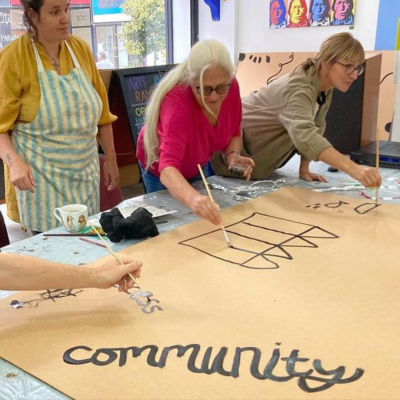 A group of people standing around a table. They are leaning over, painting with black paint onto a piece of cardboard. The word 'community' has been painted by someone at the bottom of the cardboard.